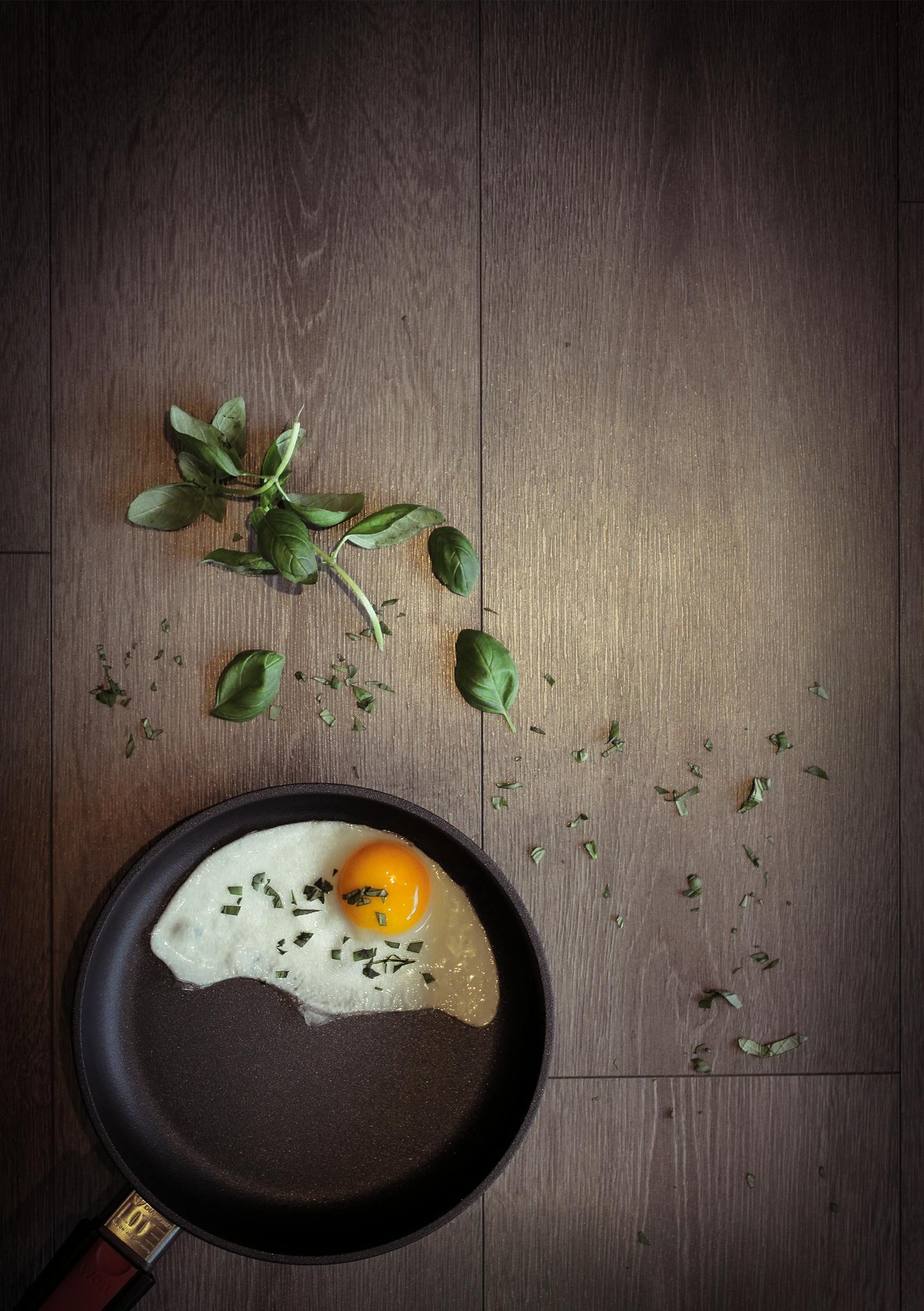 Overhead shot of a fried egg in a pan with fresh basil leaves on wooden floor.
